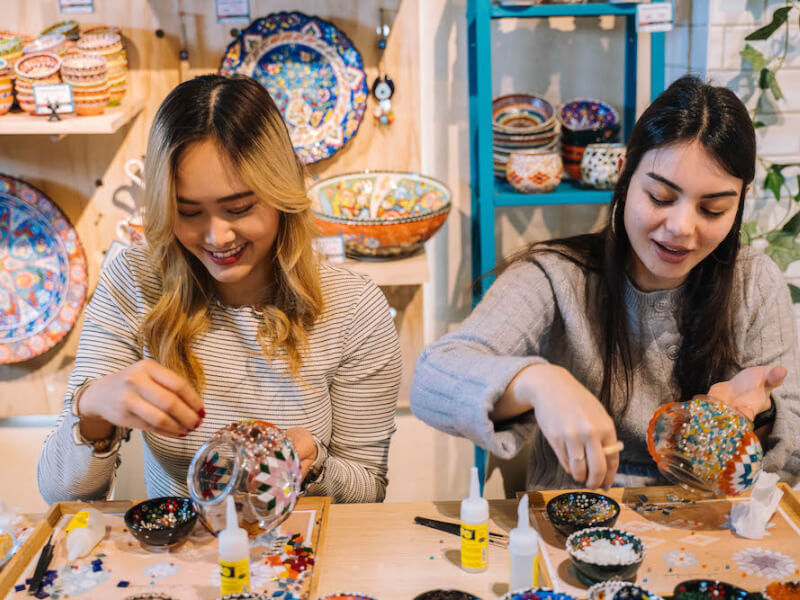 Two women smile as they make mosaic lamps